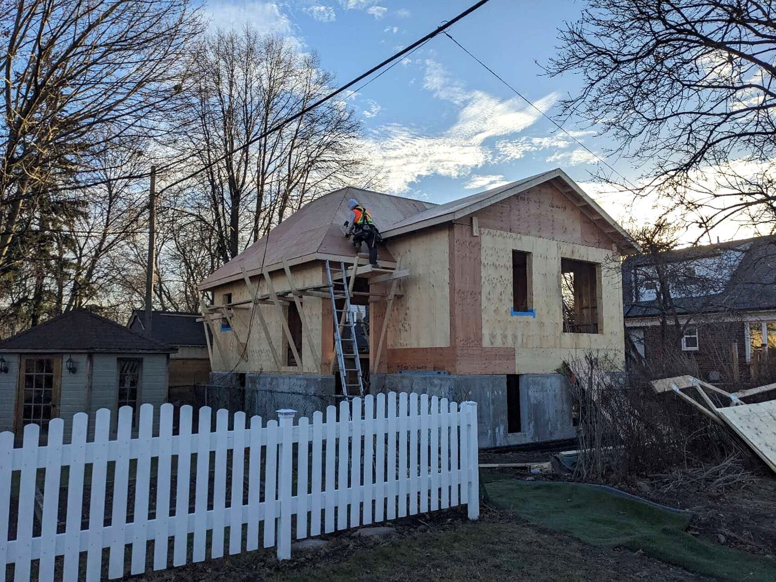 Laneway house under construction behind a white picket fence in evening light
